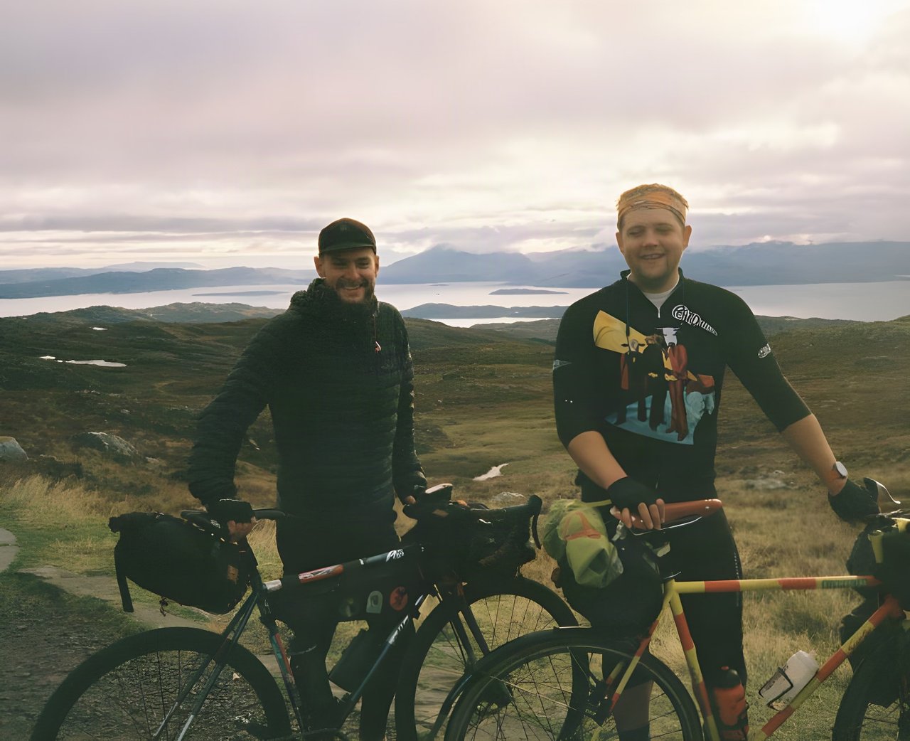 Two smiling riders with loaded touring bikes on a hilltop above a sea loch on a fixed-gear tour of north-west Scotland.