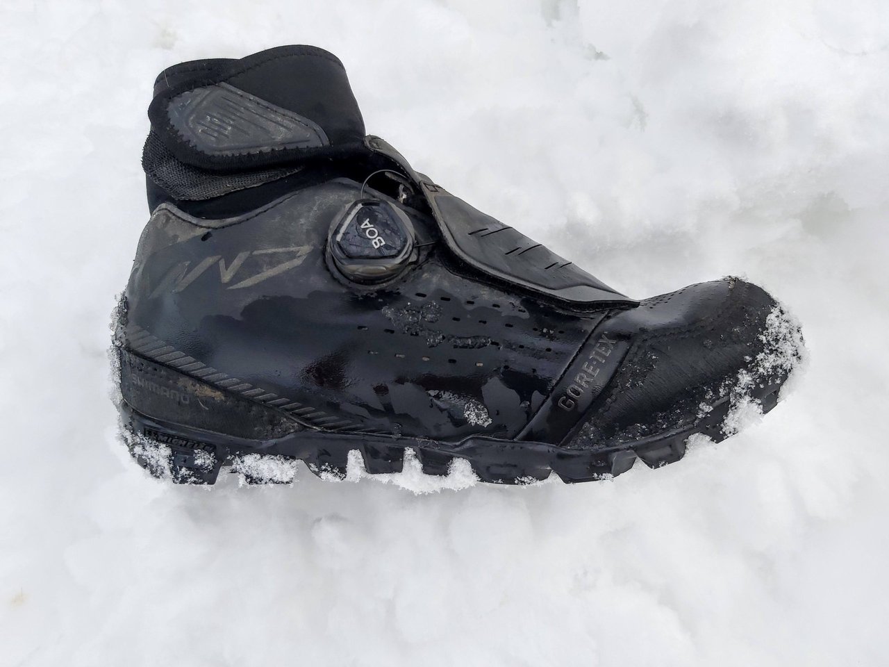 Black Shimano MW7 winter MTB shoe with BOA dial and Gore-Tex logo, resting in fresh snow.