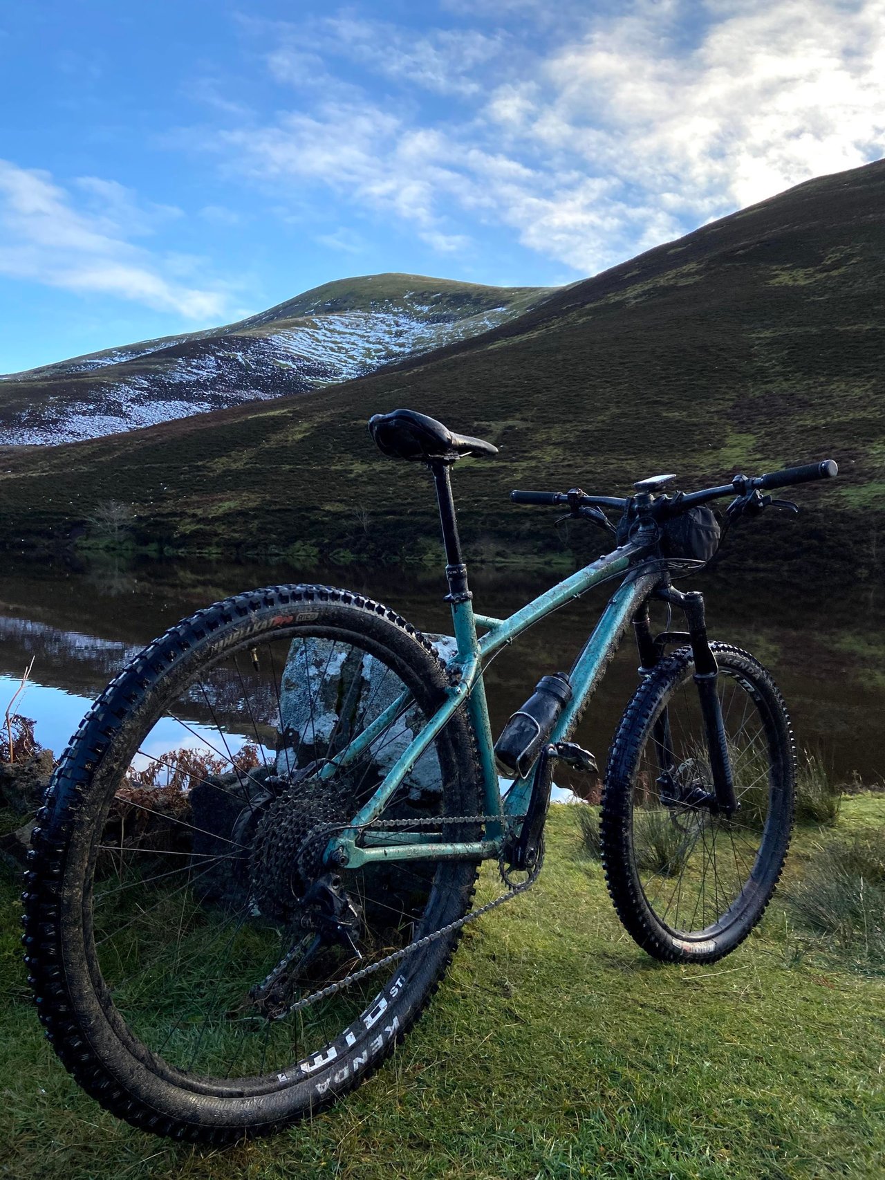 Bombtrack Cale AL hardtail in mint green stands beside a still reservoir below the snow-dusted Pentland Hills.