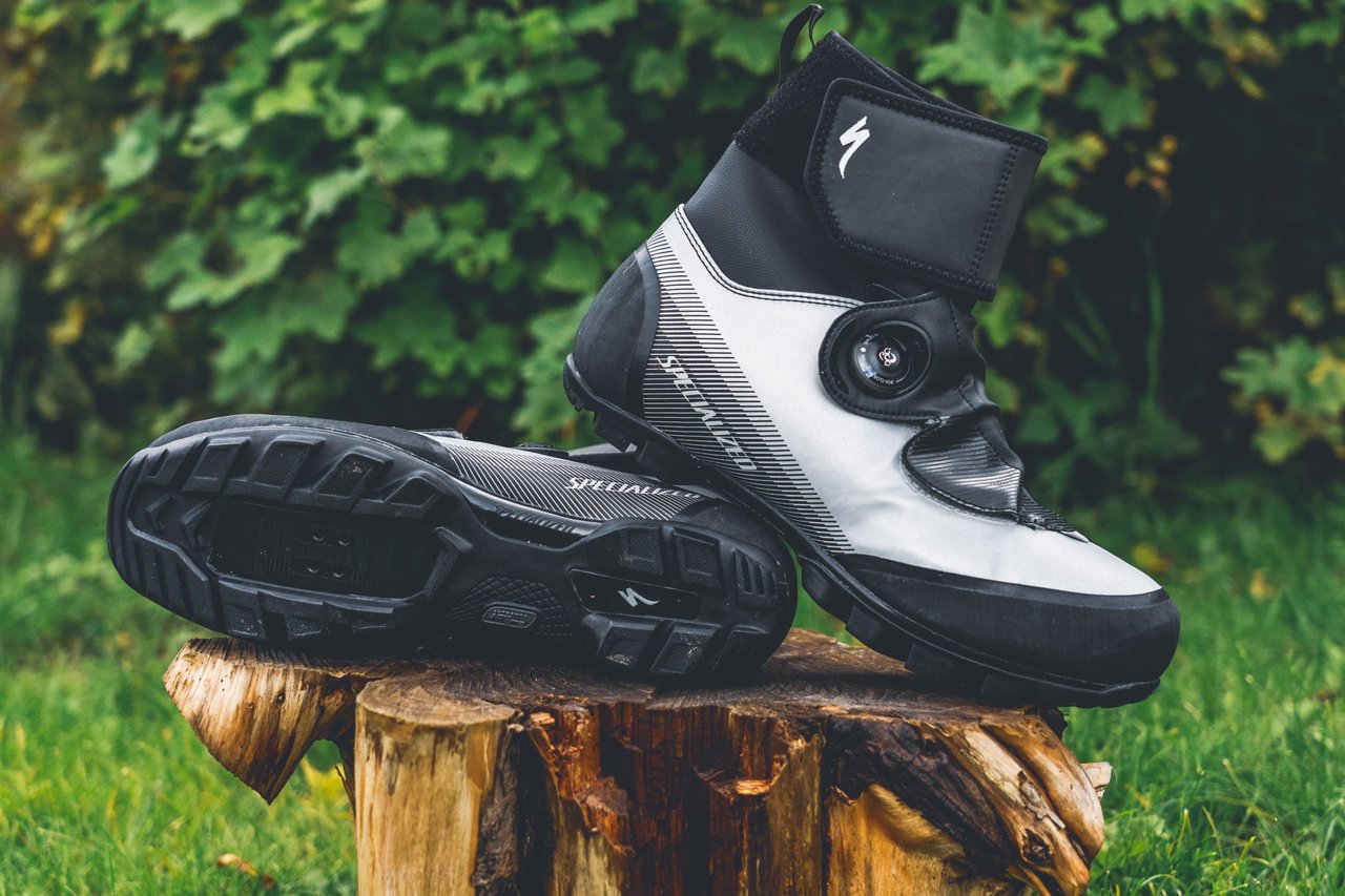 Specialized Defroster Trail MTB winter boots in reflective silver and black, one on its side showing the sole, on a log.