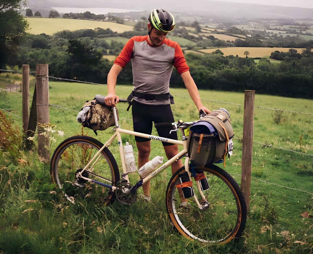 Rider with a vintage Specialized Rockhopper mountain bike loaded with bikepacking bags beside a fence in farmland.