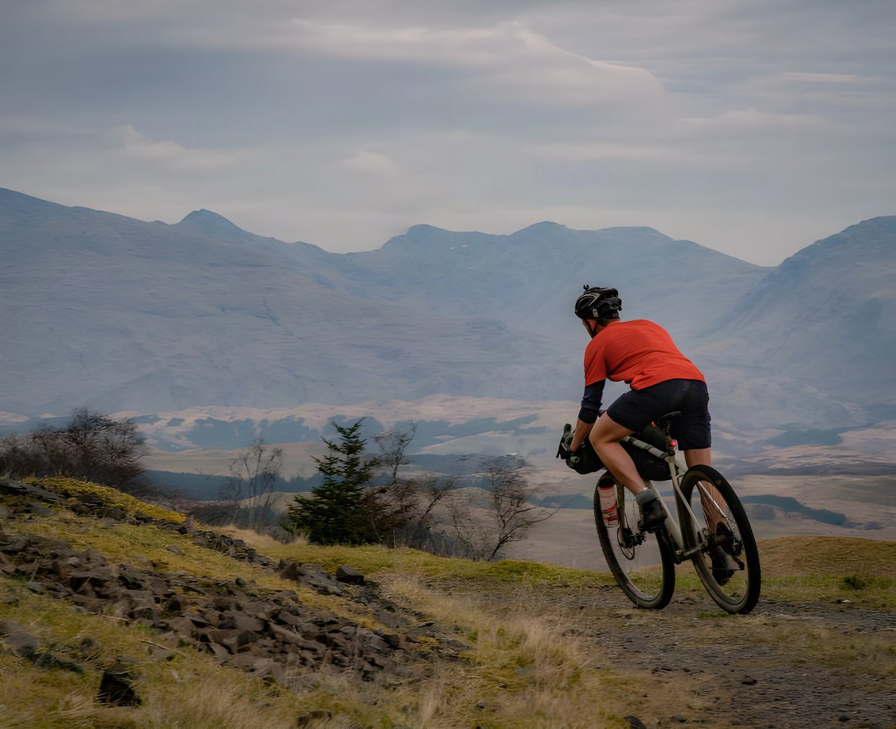 Rider in an orange top rides a rocky gravel track with Argyll hills behind, on a bikepacking overnighter.