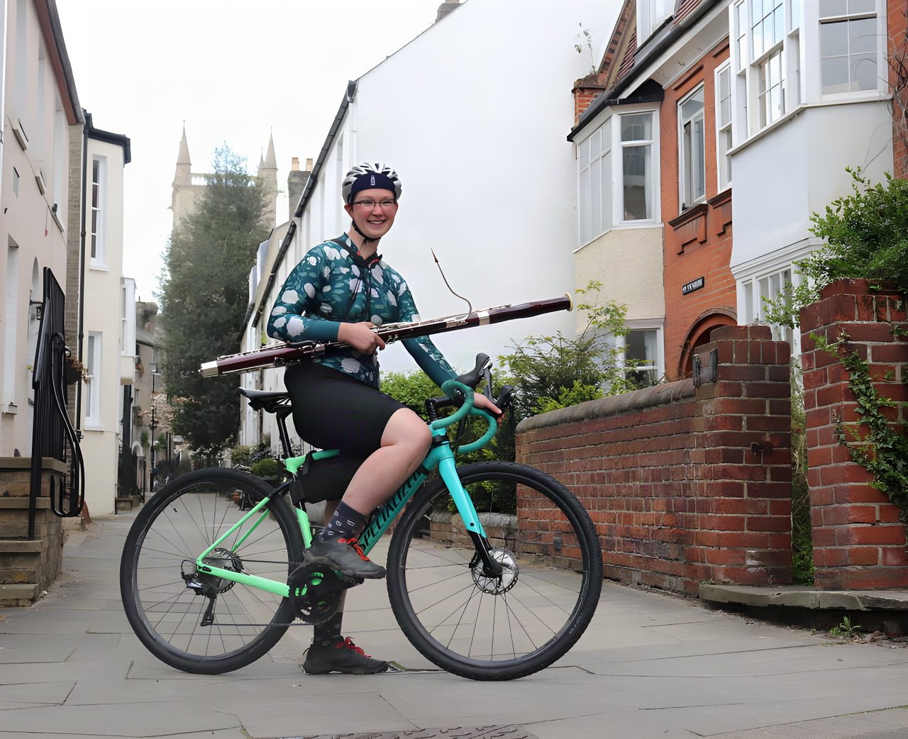 Bassoonist Alison Wormell sits astride her mint green Specialized bike on a town street, holding her bassoon.