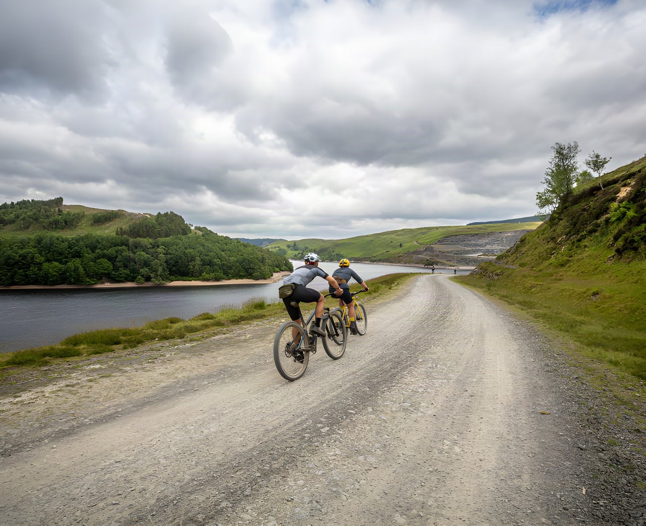 Two Gritfest riders on a wide gravel track beside a reservoir under cloudy skies in the Cambrian Mountains.