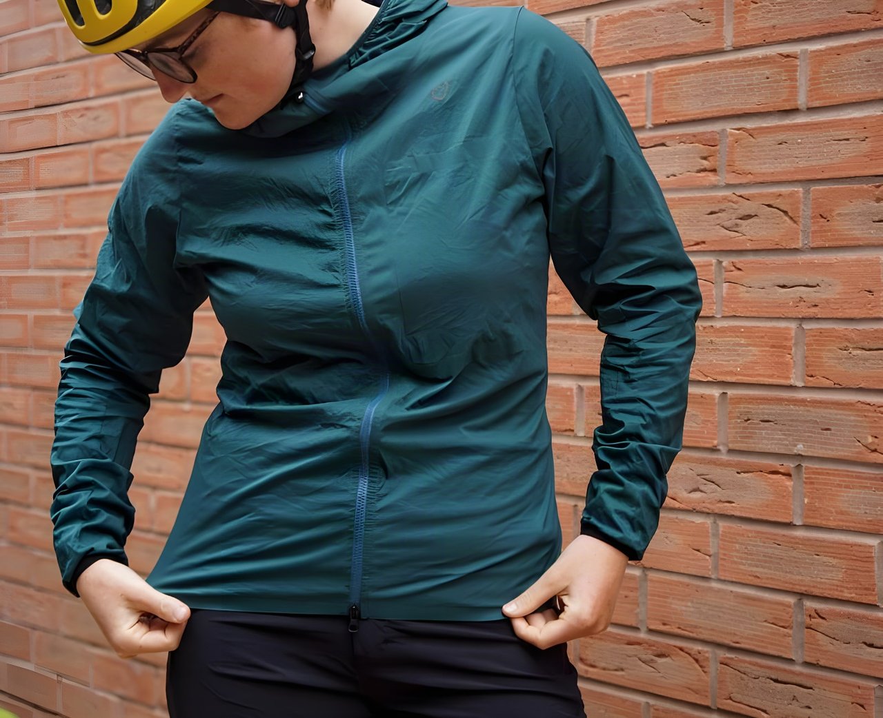 Velocio Ultralight Hooded Jacket in the deep sea colourway, worn with a yellow helmet against a brick wall.