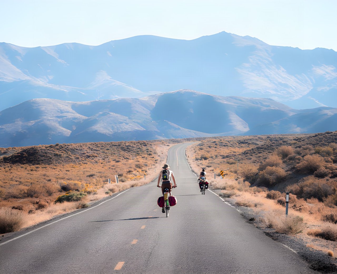 Two loaded touring cyclists ride a straight desert road towards mountains, from the Cycling Through A Pandemic book.