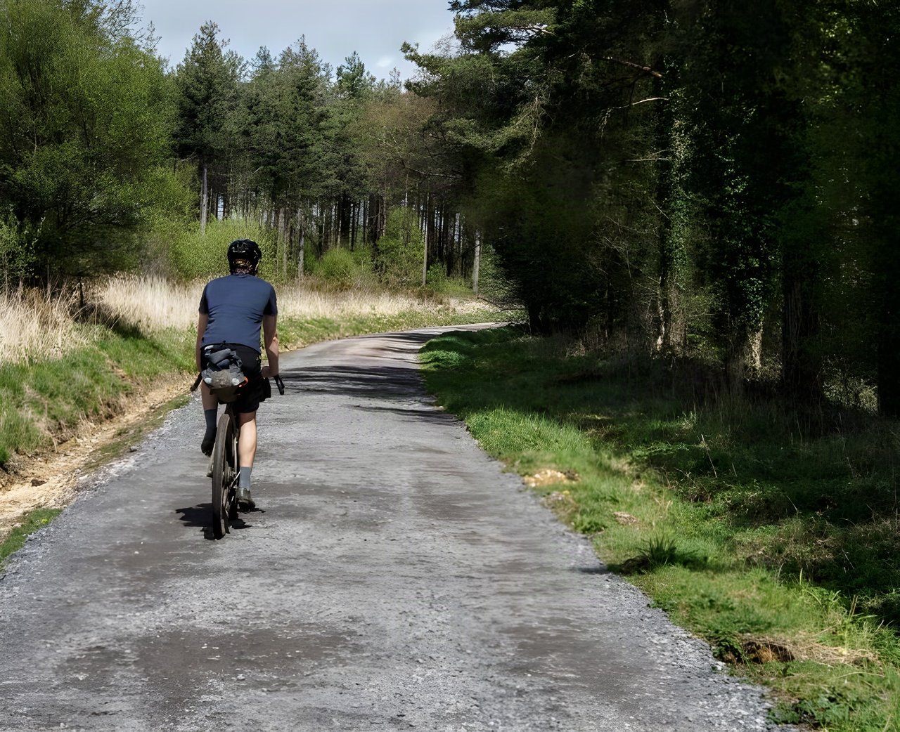 Rider in Chapeau gravel kit with a handlebar bag pedals away along a tree-lined gravel track.