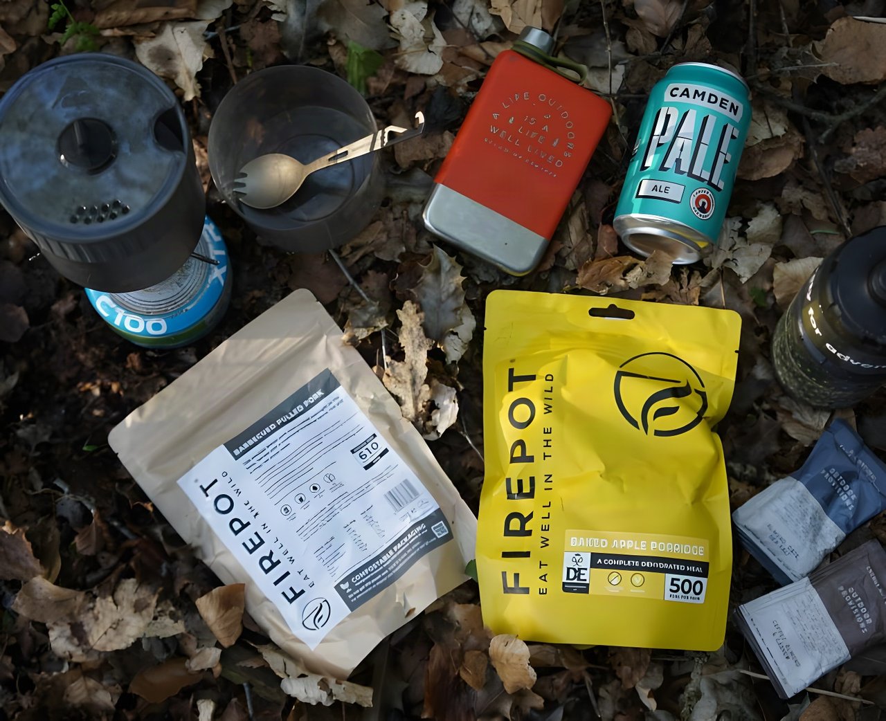 Firepot dehydrated meal packets laid out in leaf litter with a camping stove, spork, flask and a can of beer.