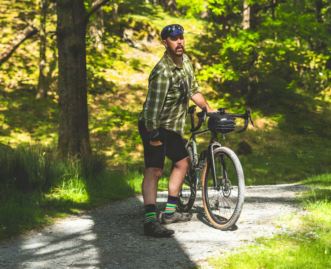 Rider wearing Lusso Adventure bib shorts stands beside a gravel bike with a bar bag on a woodland track.
