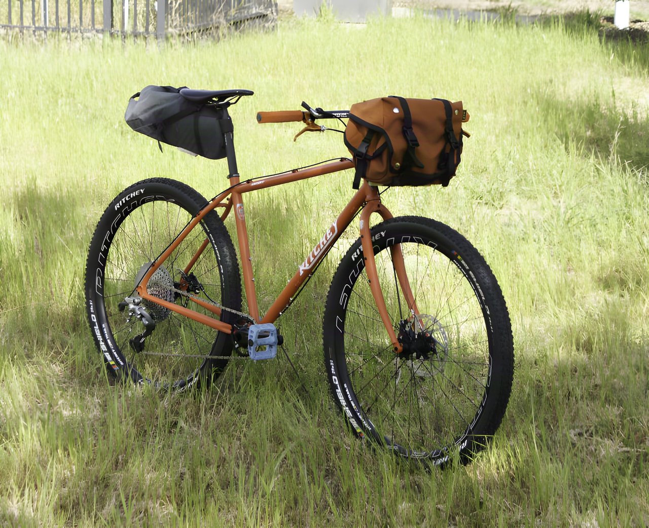 A loaded Ritchey Ascent bike with a saddle bag and handlebar bag parked in long grass.