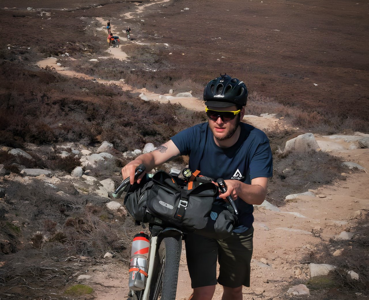Rider in Vulpine gravel shorts pushes a bikepacking bike with a handlebar bag up a rocky heather track in Scotland.