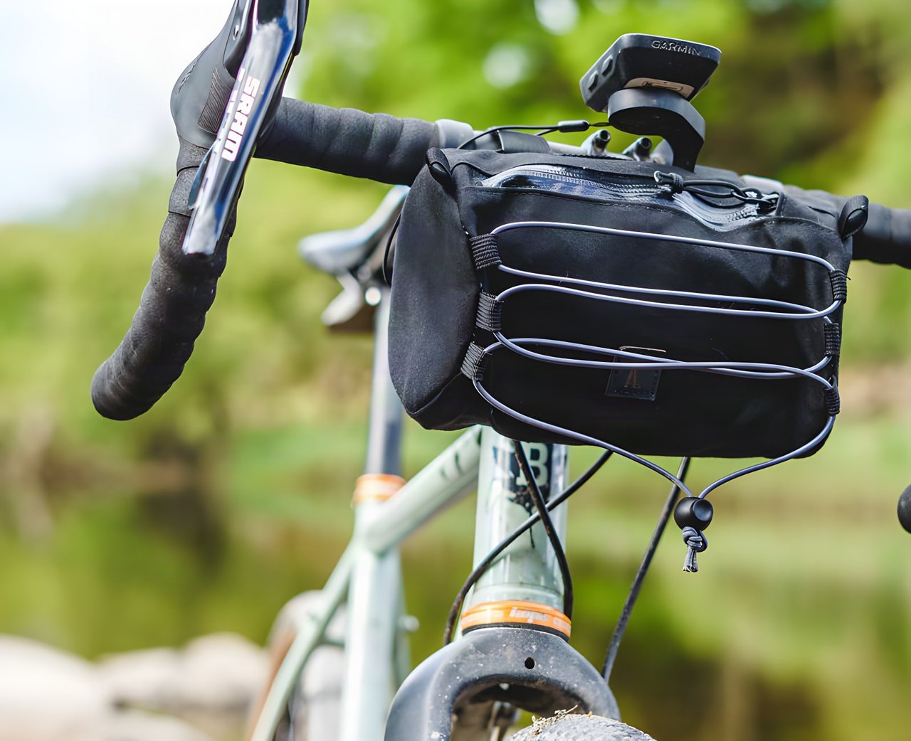 Close-up of the Ashdown Bags bar bag strapped under a gravel bike's handlebars, elastic cord on the front, beside a lake.