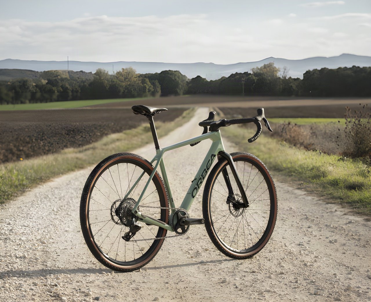 2022 Orbea Terra gravel bike in sage green parked on a white gravel track between fields, hills behind.