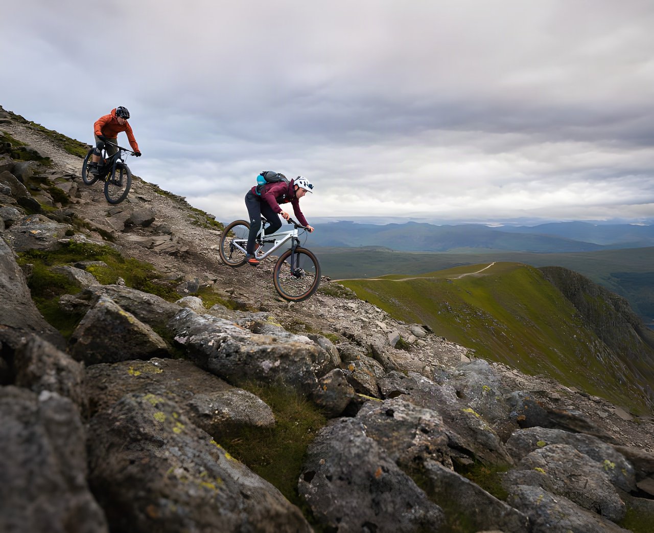 Two Rider Resilience mountain bikers descend a rocky ridge track above a green valley under heavy cloud.