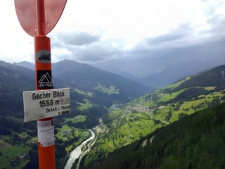 Gacher Blick 1559 m sign on an orange post above a green Alpine valley under grey cloud, on the Transalp route.