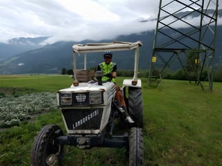 Cyclist in a hi-vis gilet sits on an old Lamborghini tractor in a field below cloud-covered Alpine slopes.