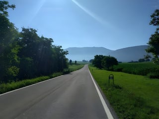 Empty tarmac road runs straight through green fields towards hazy hills in the early morning light.