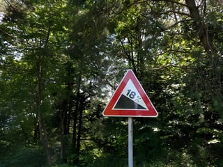 Red-bordered road sign warning of an 18 per cent descent, set among dense green trees.