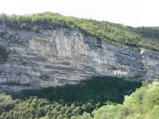 Small white building set into a ledge of a sheer limestone cliff above wooded slopes in northern Italy.