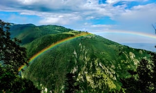 Rainbow arcs over a steep forested Italian mountainside under a mix of blue sky and cloud.