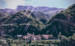 Small Italian village of terracotta-roofed houses at the foot of steep wooded mountains, with sunbeams across the slopes.