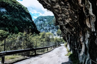 Narrow road with a guardrail runs beneath an overhanging rock face through a green gorge in the Italian Alps.
