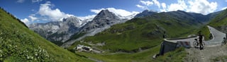 Panorama of a rider with a road bike at a hairpin high on an Alpine pass, with snowy peaks and a hotel in the valley below.