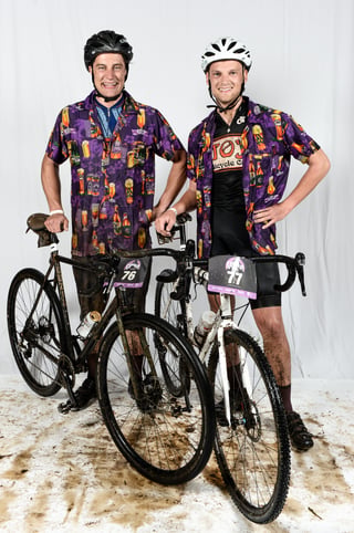 Two muddy Grinduro Scotland riders in matching beer-print shirts pose with their bikes, numbers 76 and 77.