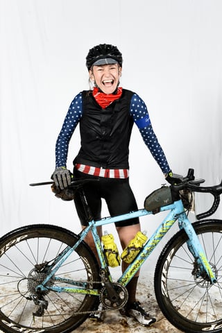 Smiling Grinduro Scotland finisher in a stars-and-stripes jersey stands with a muddy light blue gravel bike.
