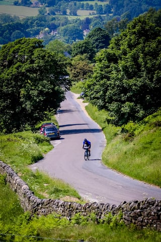 Rider climbs a winding Peak District lane between stone walls and trees, with parked cars at the roadside.