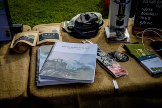 Pannier stall table with Ember coffee, a cap, an AeroPress, an Exped dry bag and a Rough Stuff Cycling in the Alps book.