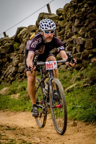 Rider with number 001 on a gravel bike rattles down a stony trail past a dry stone wall at Yorkshire True Grit.