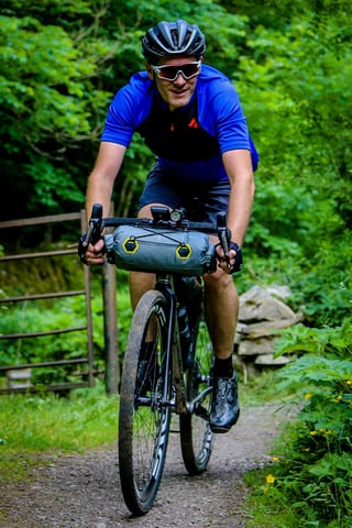 Rider with an Apidura handlebar bag and front light rides a gravel track past a metal gate on The Distance.