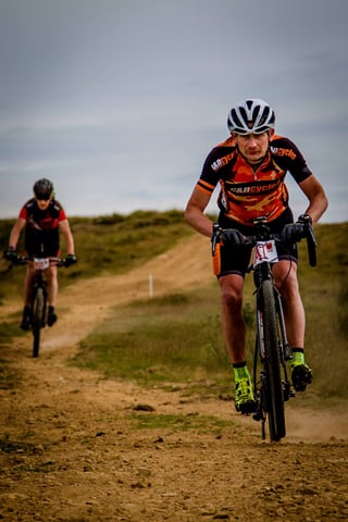 Rider in a camouflage-orange C&amp;N Cycles jersey climbs a dirt track with another rider behind at Yorkshire True Grit.