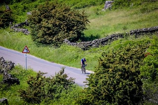 Distant rider takes a bend in a Peak District road between dry-stone walls and bushes on The Distance.
