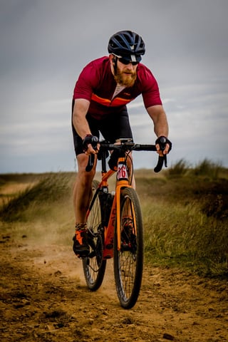 Bearded rider in a maroon jersey on an orange gravel bike rides a dusty moorland track at Yorkshire True Grit.