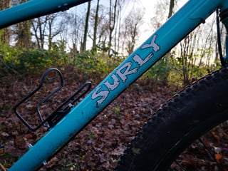 Surly logo on the teal down tube of the Bridge Club, next to a bottle cage and the front tyre.