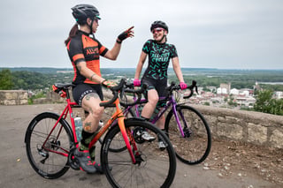 Two All-City riders with Zig Zag bikes pause and chat at a hilltop viewpoint above a river town.
