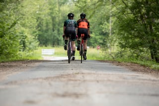 Rear view of two All-City riders on Zig Zag bikes heading away along a tree-lined gravel-edged road.