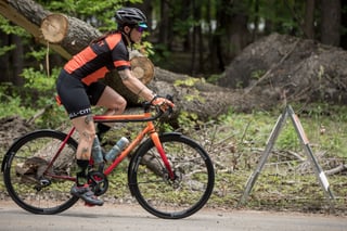Rider in All-City kit rides the orange-red fade Zig Zag with mudguards past felled tree trunks, side view.