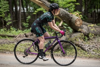 Rider in black and teal All-City Cycles kit rides the purple-lilac Zig Zag past a fallen tree trunk, side view.