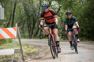 Two riders on All-City Zig Zag road bikes round a bend on a shaded gravel-edged lane past a striped road barrier.