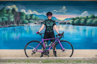 Smiling rider stands behind a purple-lilac All-City Zig Zag in front of a painted river mural.
