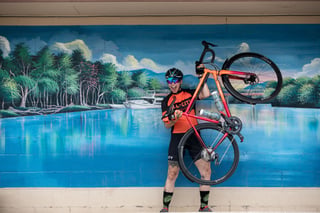 Rider in All-City kit hoists an orange-red Zig Zag overhead in front of a painted lakeside mural.