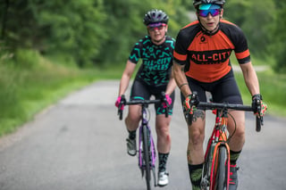 Two All-City riders climb a gentle rise on Zig Zag bikes, one in orange kit and one in black and teal, between green verges.