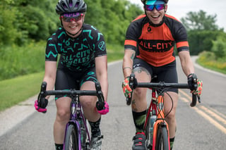 Two laughing riders in All-City Cycles kit ride side by side on Zig Zag bikes along a quiet tree-lined road.