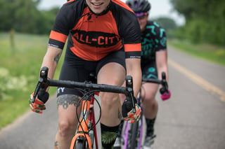 Smiling rider in an All-City jersey on a Zig Zag, close on the handlebars, with a second rider behind on a country road.