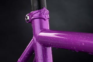 Seat collar and top tube junction of a purple All-City Zig Zag frame, with the cast A.C. collar beaded with water.