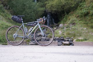 Trek Crockett 7 loaded with seat pack and frame bag, standing on a gravel track in front of a rocky stream.
