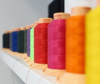 Reels of coloured sewing thread on a shelf in the Straight Cut studio, from dark blue and red to pink, orange and yellow.